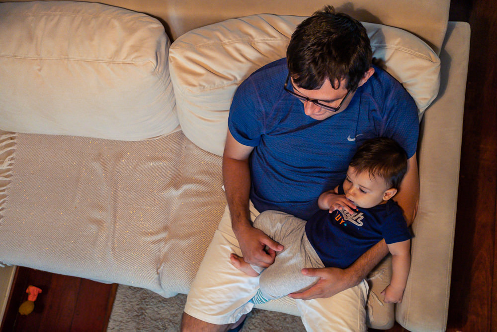 Fotografia Documental de Família. Foto da rotina da família com seu filho criança bebê, mostrando a essência a história da família.  Foto em Barueri, SP. Criança no chiqueirinho cercado com TV ao fundo assistindo video do Pedrinho e Sofia do TV JW 
