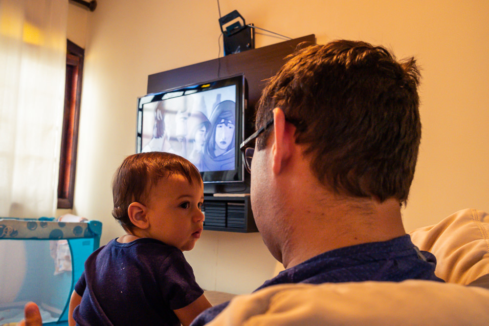 Fotografia Documental de Família. Foto da rotina da família com seu filho criança bebê, mostrando a essência a história da família.  Foto em Barueri, SP. Criança no colo do papai com TV ao fundo assistindo video do Pedrinho e Sofia do TV JW 