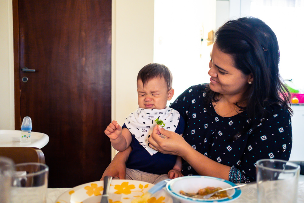 Fotografia Documental de Família. Foto da rotina da família com seu filho criança bebê, mostrando a essência a história da família.  Foto em Barueri, SP. Mãe alimenta seu filho com alface que faz careta demonstrando que não gosta