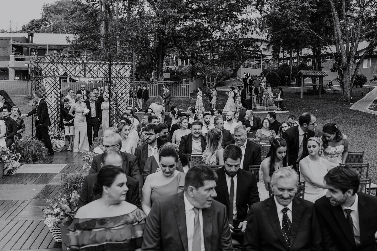 um lindo casamento no campo, mostra todos os convidados posicionado nas cadeiras esperando para o inicio das cerimonia. o fotografo daniel okuyama busca retratar as expressões das pessoas emocionadas.