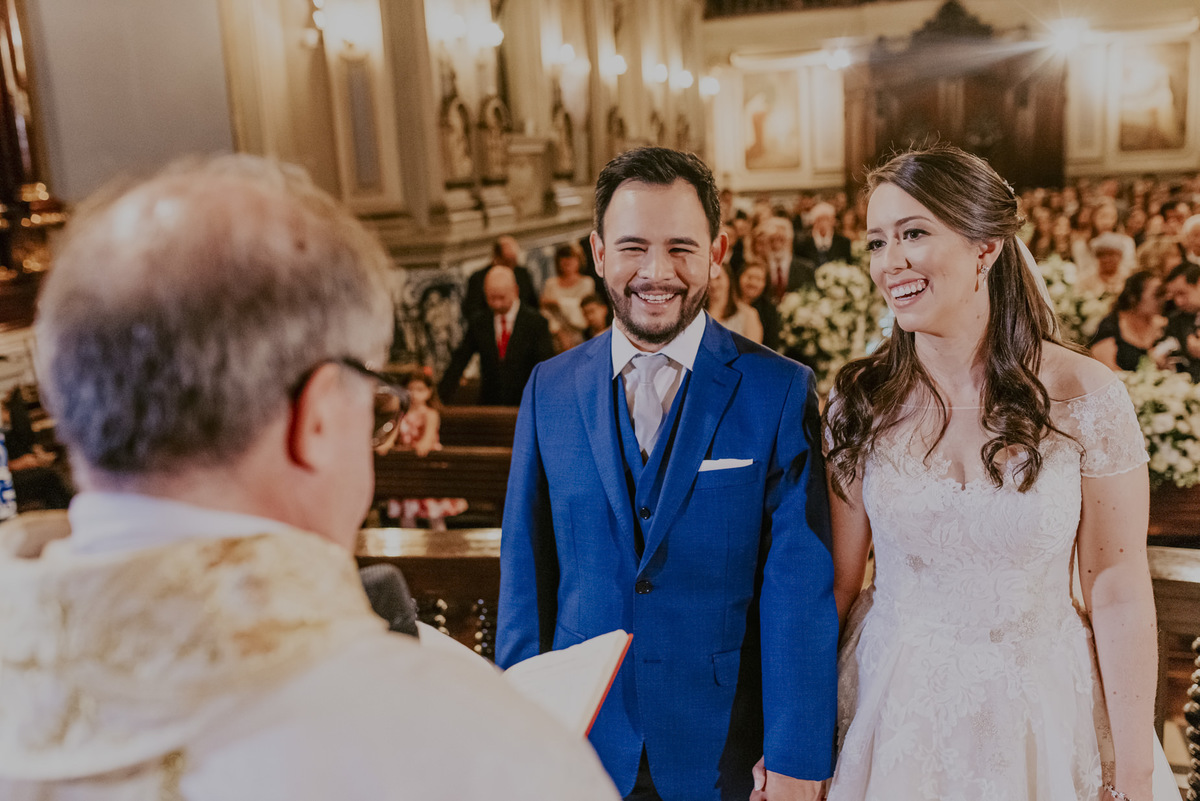 o padre com muito carinho celebra o matrimonio dos noivos com muita alegria e amor. o sorriso dos noivos com todo esse carinho na capela da puc sp