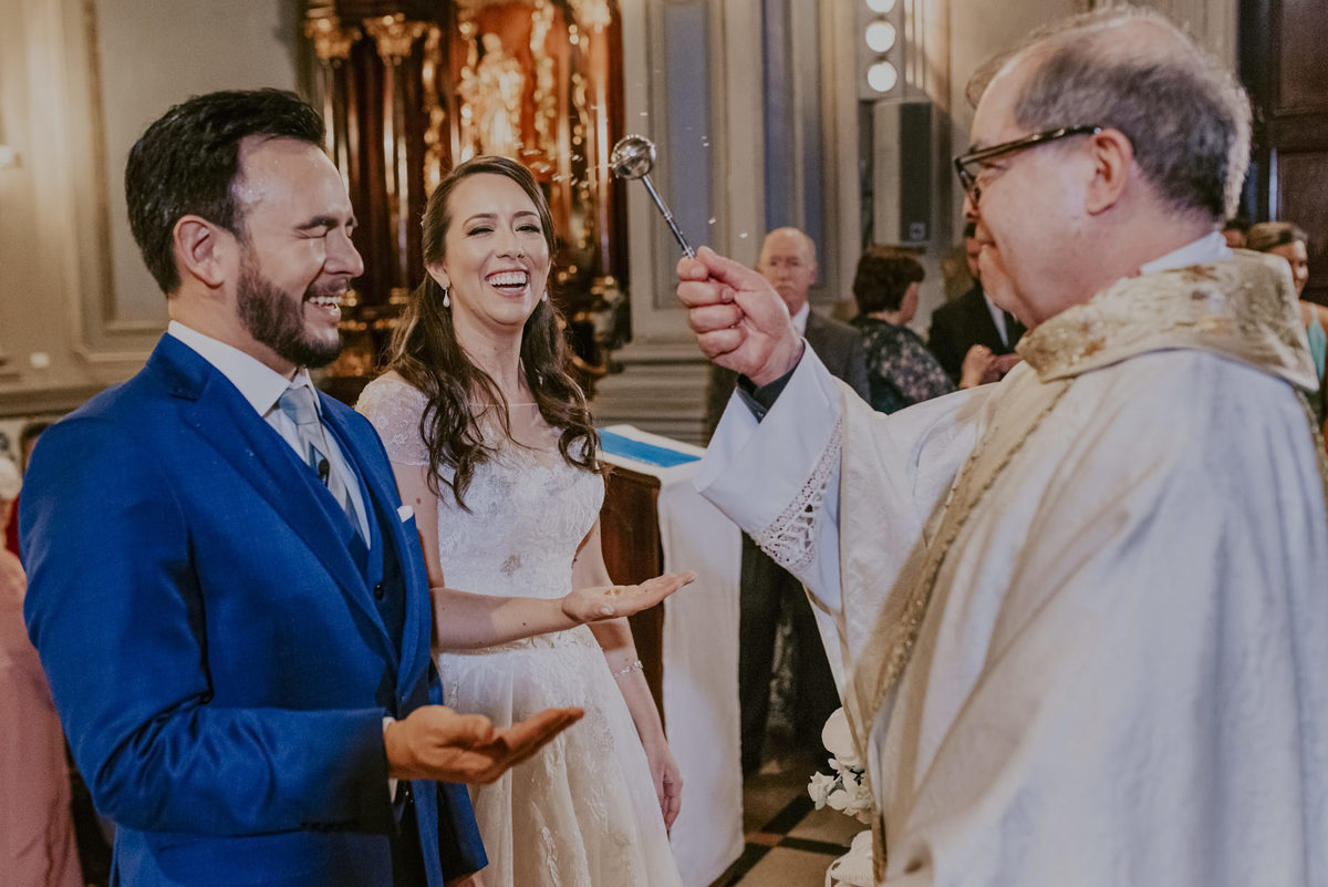o padre da capela da puc ao benzer os noivos joga agua benta no noivo que fecha os olhos e a noiva sorri, momento decisivo da fotografia de casamento na igreja da puc sp foto de daniel okuyama