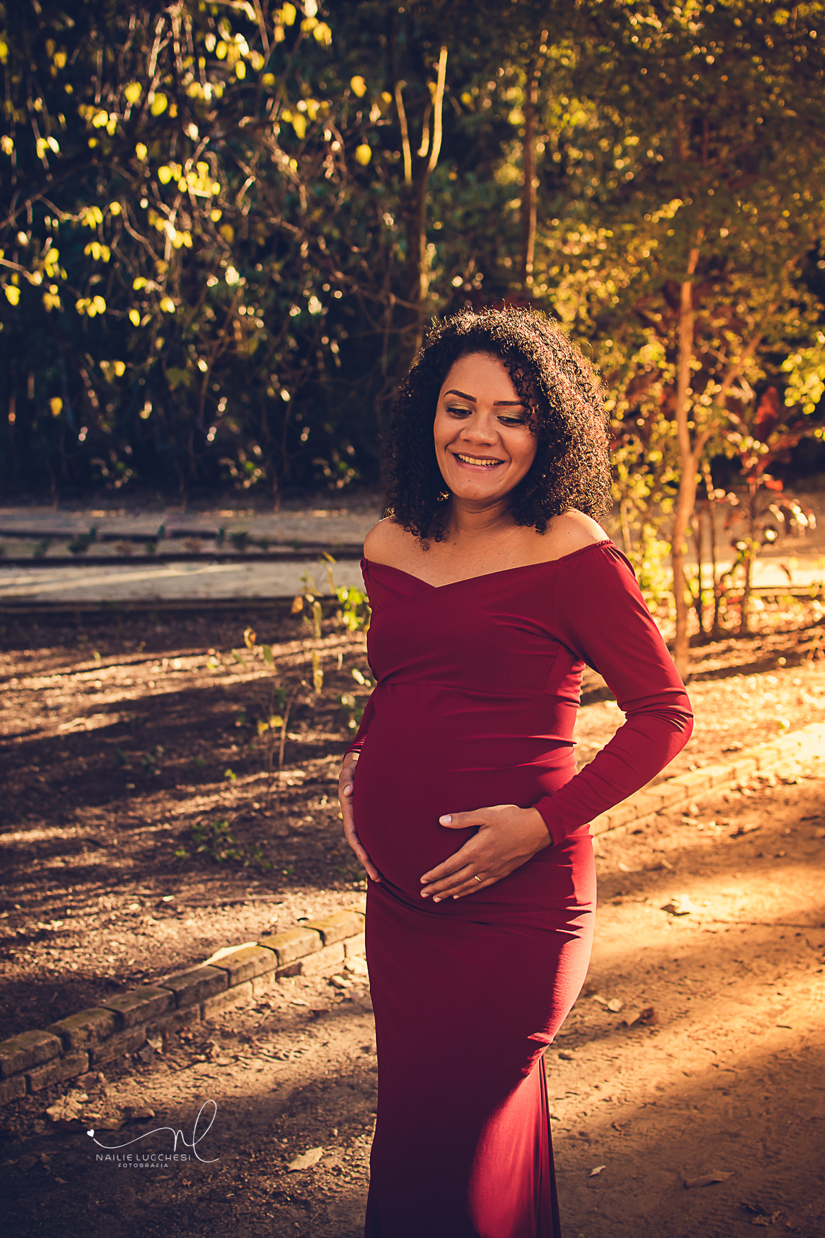 gestante linda e feliz sorrindo com as mãos na barriga no estudio amora, ela veste um lindo vestido longo e vermelho