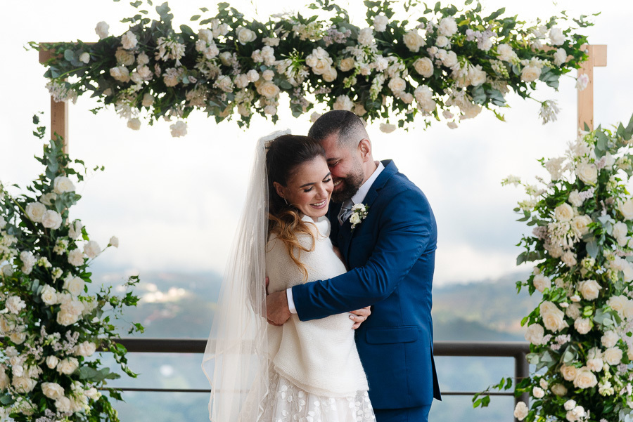 Abraço emocionado de Chico e Cacau sob o arco de flores brancas. Casamento ao ar livre no Bendito Cacao, Campos do Jordão. O cenário de montanha e o visual elegante dos noivos, com a noiva usando uma echarpe, refletem o aconchego da serra.