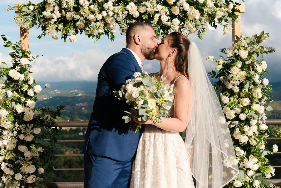Beijo apaixonado de Chico e Cacau sob o arco floral com vista para as montanhas. Casamento romântico ao ar livre no Bendito Cacao, Campos do Jordão. O registro perfeito do amor em um cenário de natureza exuberante e sofisticação na serra.