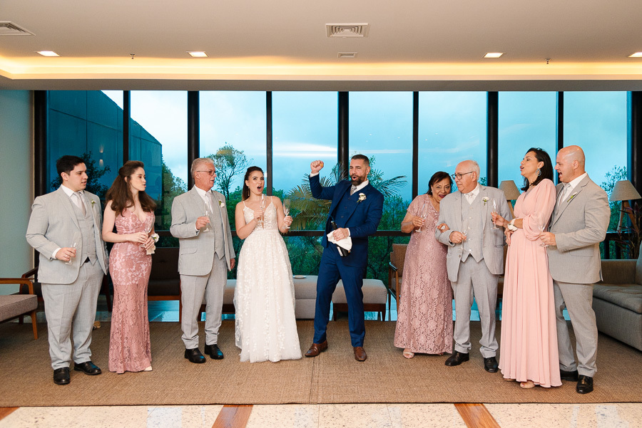 Chico e Cacau brindam com a família no Bendito Cacao. O noivo comemora vibrando ao lado dos pais e padrinhos, tendo ao fundo a vista panorâmica das montanhas de Campos do Jordão através das janelas de vidro. Celebração emocionante e sofisticada na serra.