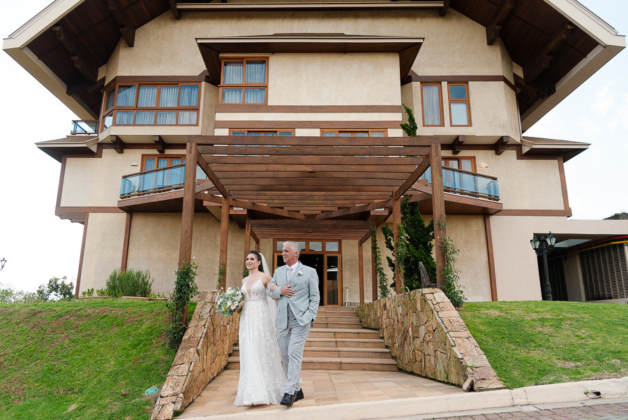 Chegada triunfal da noiva Cacau com seu pai no Bendito Cacao. A arquitetura imponente do hotel em Campos do Jordão serve de cenário para a entrada, destacando a elegância e a sofisticação deste casamento na montanha.
