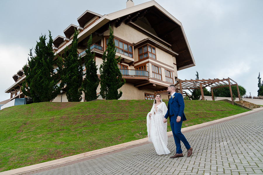 Chico e Cacau caminhando felizes em frente à fachada imponente do Bendito Cacao Resort & Spa. Ensaio de casamento externo destacando a arquitetura estilo europeu e o clima de montanha em Campos do Jordão.