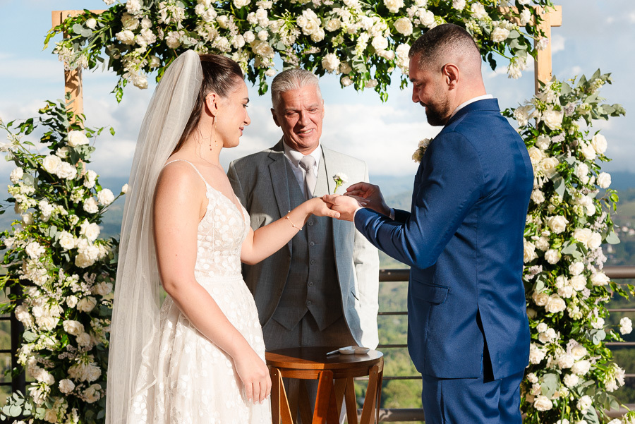 O momento do 'sim': a emocionante troca de alianças de Chico e Cacau, sob o olhar carinhoso do celebrante. Cerimônia ao ar livre com vista para as montanhas no Bendito Cacao, em Campos do Jordão. Fotografia de casamento que eterniza a conexão verdadeira.