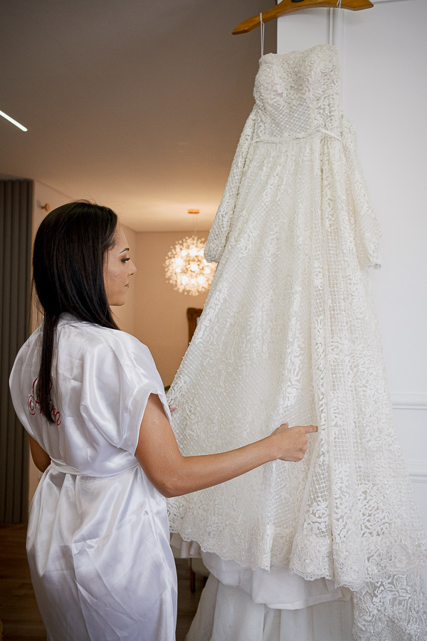 A delicadeza em cada fio. O vestido de noiva com mangas de renda iluminado pela luz suave da janela. Inspiração romântica e elegante para casamentos em São José dos Campos.
