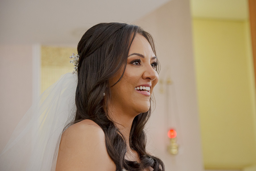 A serenidade de um sonho realizado. Close na noiva durante a bênção, destacando a maquiagem iluminada e os brincos de pérola clássicos. Inspiração de beleza para noivas.