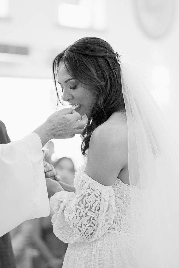 Fé e serenidade. Em preto e branco, o perfil da noiva recebendo a comunhão. O véu e o decote ombro a ombro compõem um visual angelical e respeitoso no altar.