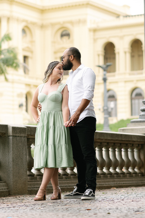Claudia e Matheus em ensaio no Museu do Ipiranga. Matheus beija a testa de Claudia, que veste um vestido verde menta. Ao fundo, a arquitetura clássica amarela do museu e uma balaustrada de pedra compõem o cenário romântico e sofisticado.