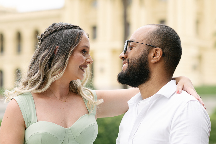 Close de Claudia e Matheus sorrindo e trocando olhares apaixonados no Museu do Ipiranga. Claudia tem a mão no ombro de Matheus, que usa óculos de sol. Ao fundo, a fachada amarela do museu aparece desfocada, destacando a cumplicidade do casal.