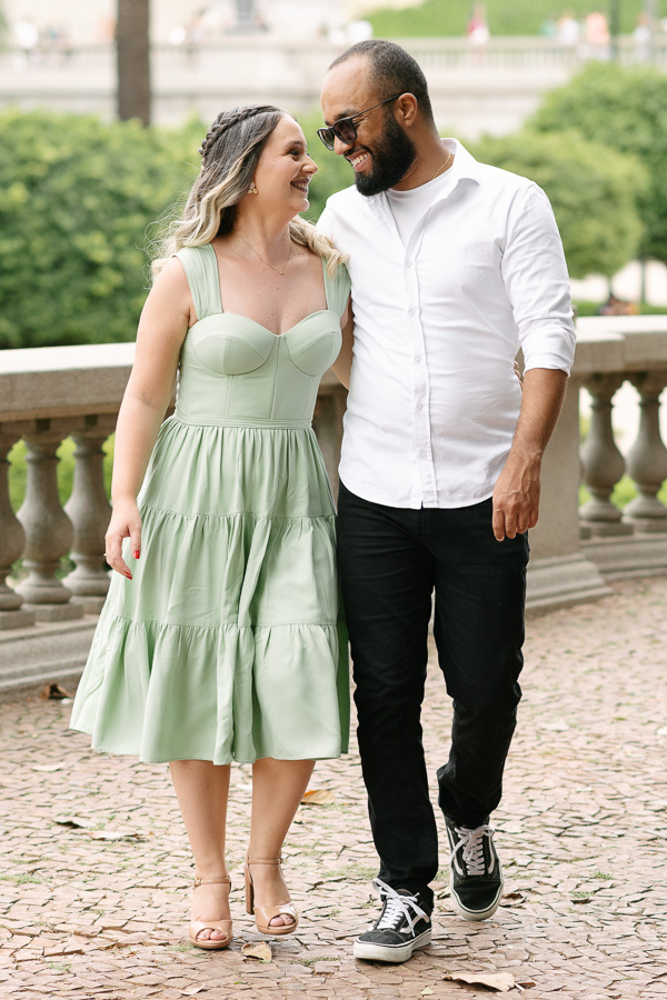 Claudia e Matheus sorriem e caminham abraçados pelos jardins do Museu do Ipiranga. Claudia usa um vestido verde menta e Matheus veste camisa branca e óculos escuros. O casal demonstra cumplicidade em um cenário histórico com muretas de pedra ao fundo.