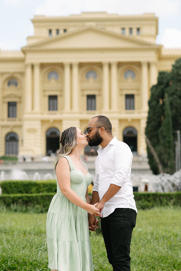 Claudia e Matheus se beijam carinhosamente em frente à imponente fachada do Museu do Ipiranga. Eles estão de mãos dadas em um gramado verde. A arquitetura neoclássica amarela do museu ao fundo completa a cena romântica e sofisticada do ensaio.