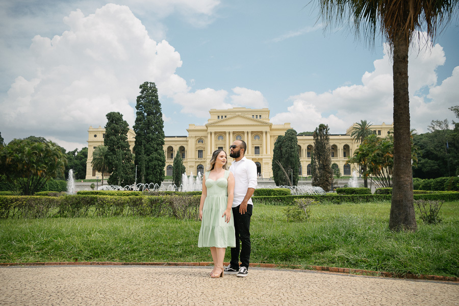 Claudia e Matheus posam no jardim do Museu do Ipiranga. Matheus está atrás de Claudia, ambos olham para o lado. Ao fundo, a imponente fachada amarela do museu, fontes de água e árvores altas sob um céu com nuvens brancas.