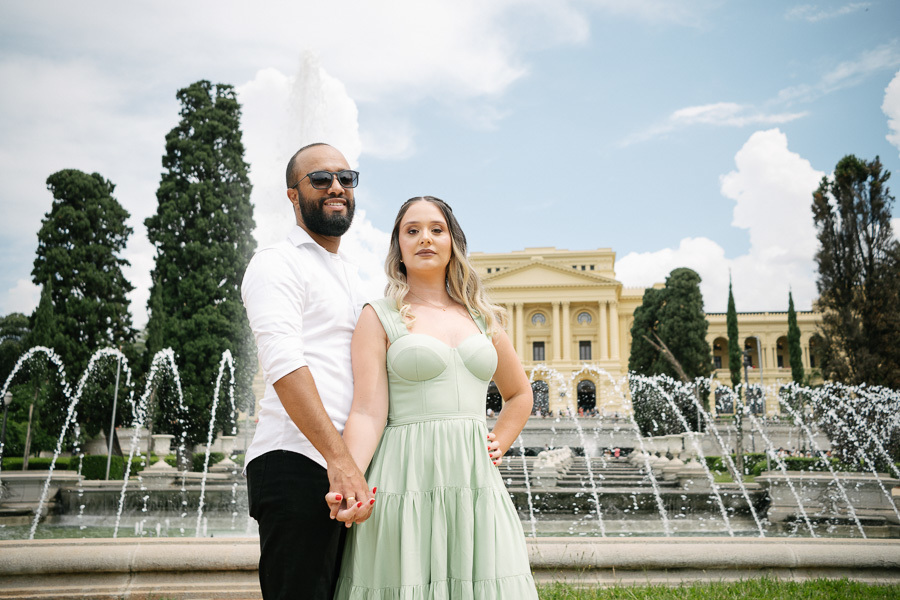 Claudia e Matheus posam de mãos dadas em frente à fonte do Museu do Ipiranga. Matheus usa óculos escuros e camisa branca; Claudia veste um vestido verde menta. A água da fonte em movimento e a fachada amarela do museu completam o cenário vibrante.