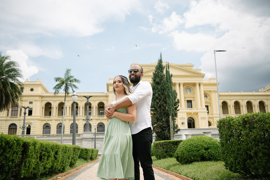 Claudia e Matheus posam de mãos dadas em frente à fonte do Museu do Ipiranga. Matheus usa óculos escuros e camisa branca; Claudia veste um vestido verde menta. A água da fonte em movimento e a fachada amarela do museu completam o cenário vibrante.