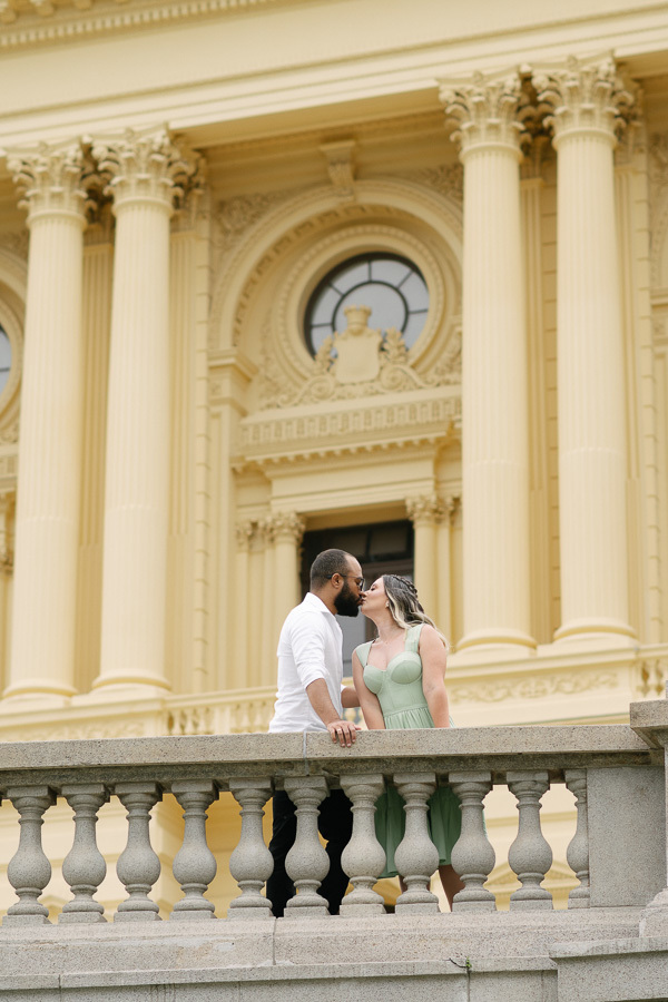 Claudia e Matheus se beijam no Museu do Ipiranga, posicionados atrás de uma balaustrada de pedra cinza. Ao fundo, as imponentes colunas clássicas e detalhes arquitetônicos da fachada amarela do museu criam um cenário monumental e romântico.