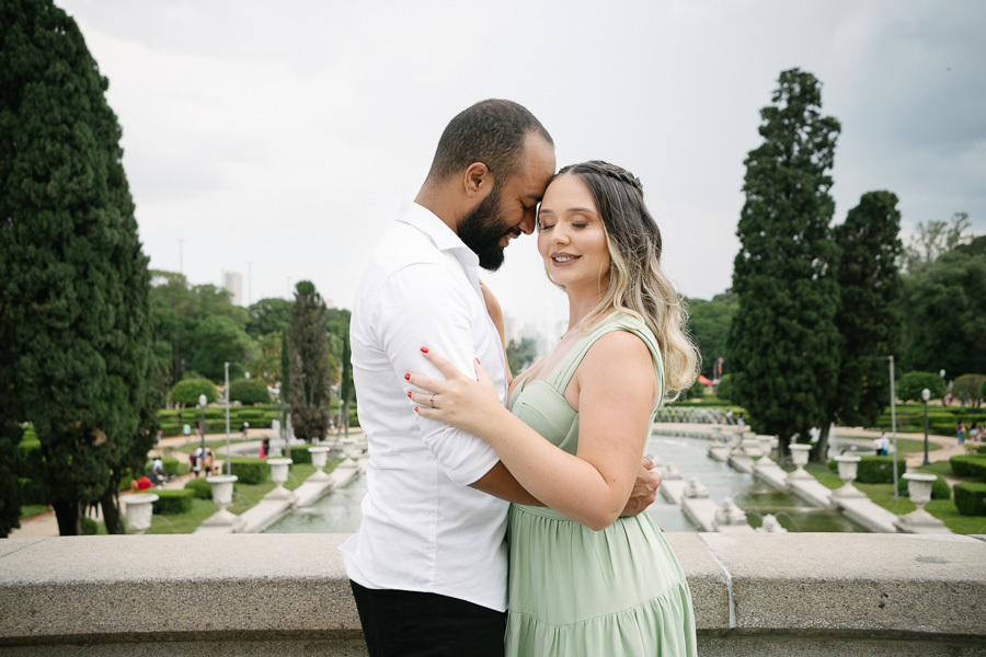 Claudia e Matheus estão abraçados com as testas encostadas em um momento íntimo no Museu do Ipiranga. Ela veste verde menta e ele camisa branca. Ao fundo, jardins simétricos, fontes de água e árvores altas completam o cenário sob um céu claro.