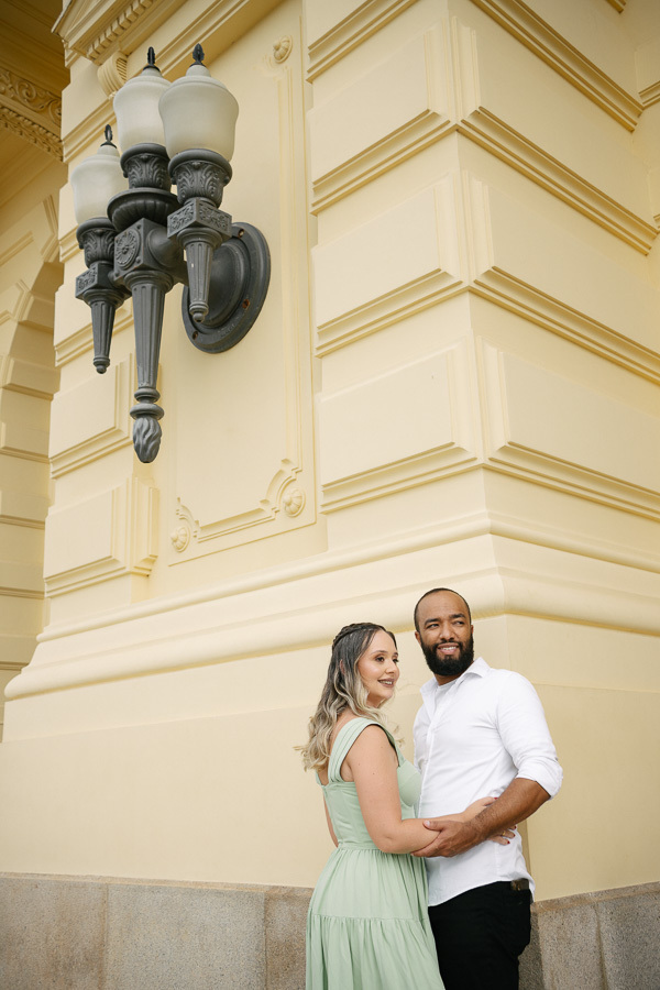 Claudia e Matheus sorriem abraçados ao lado de uma luminária clássica na fachada do Museu do Ipiranga. Claudia veste verde menta e Matheus camisa branca. A arquitetura amarela imponente destaca a sofisticação do ensaio pré-casamento.