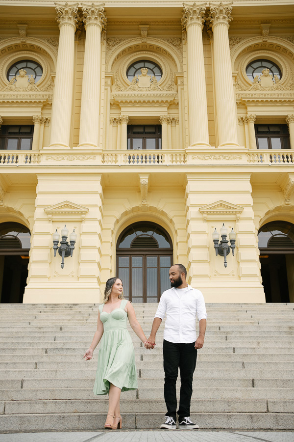 Claudia e Matheus descem de mãos dadas a imponente escadaria de pedra do Museu do Ipiranga. Claudia veste um vestido verde menta e Matheus camisa branca. Ao fundo, a grandiosa fachada neoclássica amarela com colunas e arcos destaca a elegância do casal.