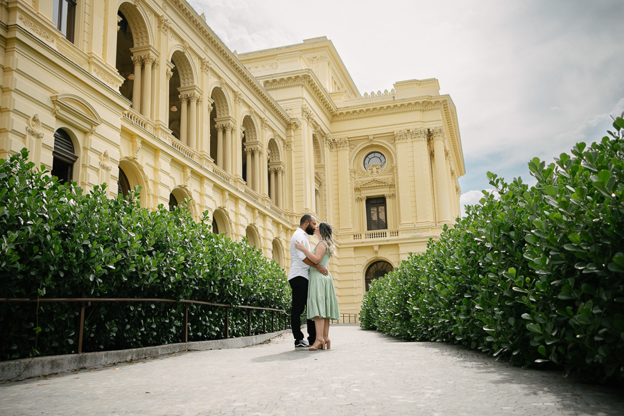Claudia e Matheus se beijam em uma passarela de pedras no Museu do Ipiranga. Eles estão emoldurados por arbustos verdes altos em ambos os lados, com a imponente arquitetura amarela e arcos do museu preenchendo o fundo sob um céu nublado.