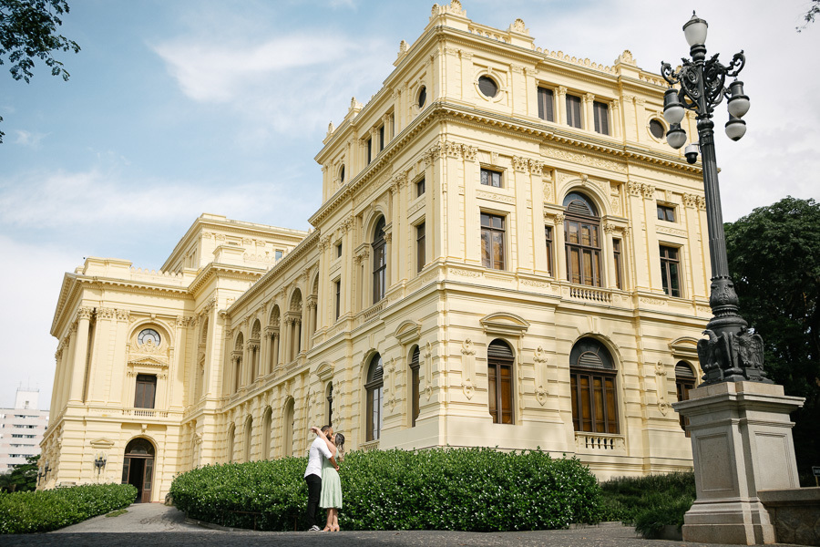 Claudia e Matheus se abraçam e se beijam em frente à lateral do Museu do Ipiranga. O casal está em uma área pavimentada, com arbustos verdes ao redor e um poste de luz clássico à direita. A arquitetura amarela do museu domina o cenário sob o céu azul.