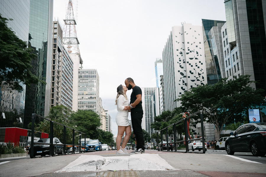 Claudia e Matheus se beijam no canteiro central da Avenida Paulista. Claudia veste um conjunto branco e Matheus usa camiseta e calça pretas. Ao fundo, a perspectiva da avenida com prédios modernos, antenas e o movimento da cidade sob um céu claro.