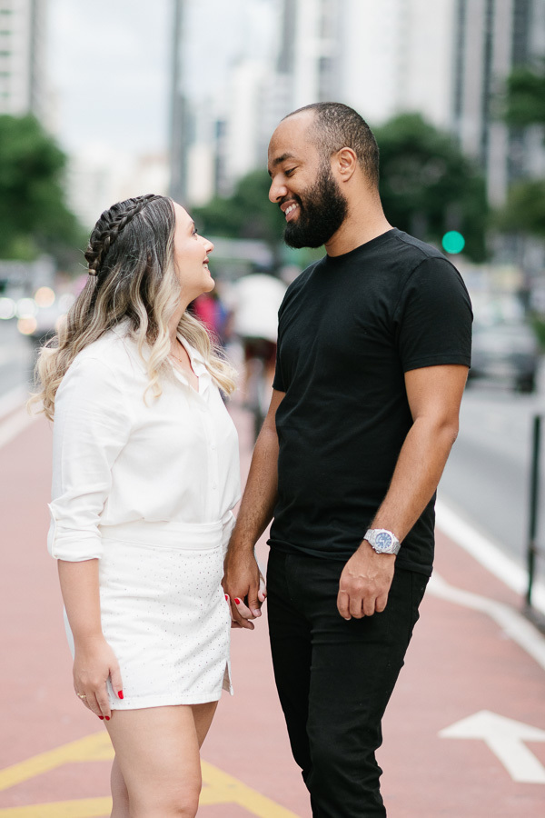 Claudia e Matheus sorriem e trocam olhares na ciclovia da Avenida Paulista. Claudia veste blusa e saia brancas, enquanto Matheus usa camiseta e calça pretas. Ao fundo, o cenário urbano com prédios e o movimento da via completam a foto pré-wedding.