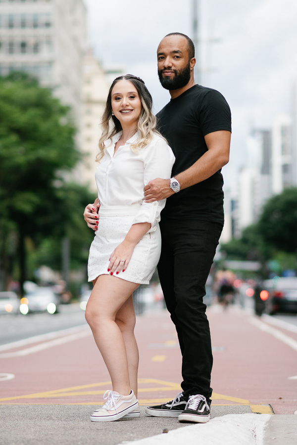 Claudia e Matheus posam abraçados na ciclovia da Avenida Paulista. Ela veste um conjunto branco e tênis; ele usa camiseta e calça pretas. Ao fundo, a via se estende com prédios modernos e luzes da cidade sob um céu nublado, em um estilo urbano.