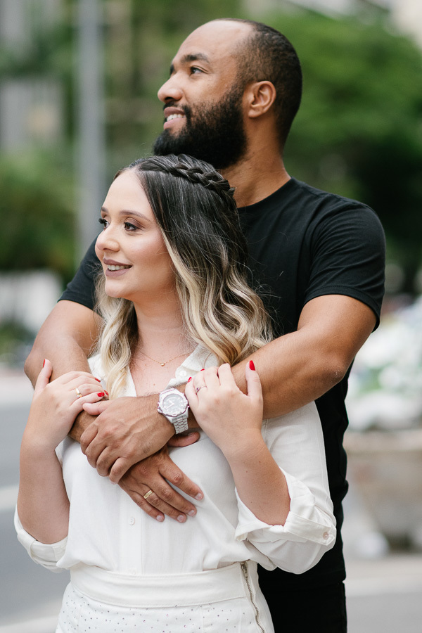 Claudia e Matheus estão abraçados e olham para o horizonte na Avenida Paulista. Matheus abraça Claudia por trás, com as mãos entrelaçadas sobre o peito dela. Ambos sorriem em um momento de tranquilidade, com o cenário urbano de São Paulo ao fundo.