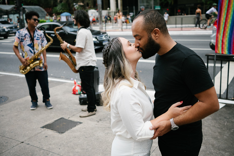 Claudia e Matheus se beijam na calçada da Avenida Paulista. Claudia veste branco e Matheus veste preto. Ao fundo, dois músicos tocam saxofone na rua, ladeados pelo movimento de carros e pedestres, trazendo uma atmosfera urbana e artística ao ensaio.