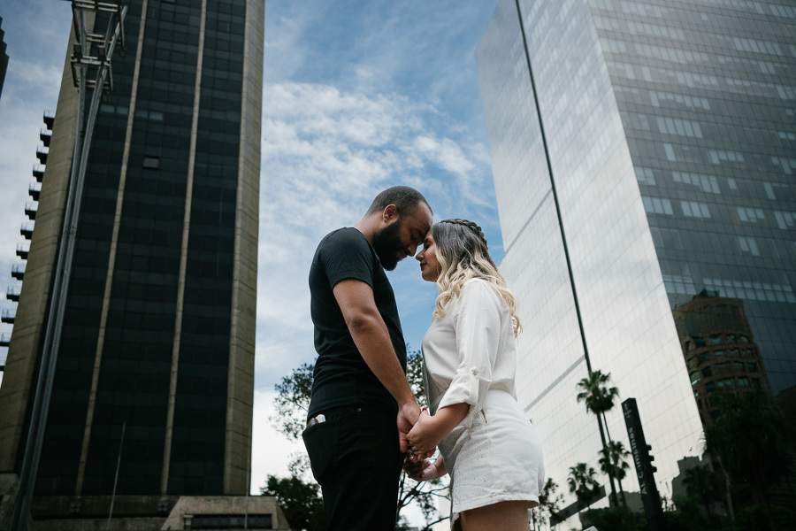 Claudia e Matheus estão de pé, frente a frente, com as testas encostadas e mãos dadas em um momento solene na Avenida Paulista. A foto, tirada de um ângulo baixo, destaca os grandes edifícios espelhados ao fundo contra um céu claro com nuvens.
