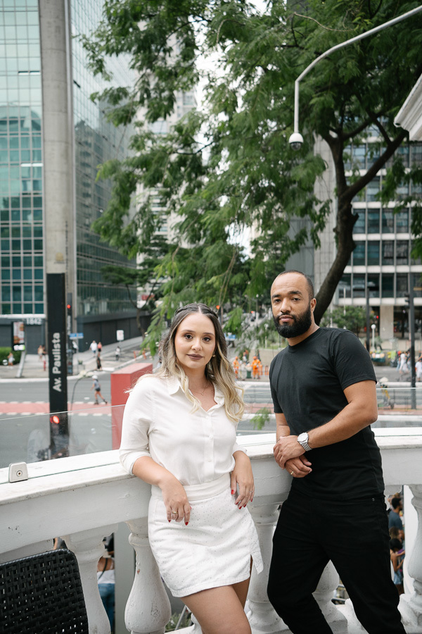 Claudia e Matheus posam em um terraço com vista para a Avenida Paulista. Claudia usa um conjunto branco e Matheus veste camiseta preta. Ao fundo, a icônica placa da avenida, árvores e edifícios modernos compõem o cenário urbano e dinâmico do ensaio.