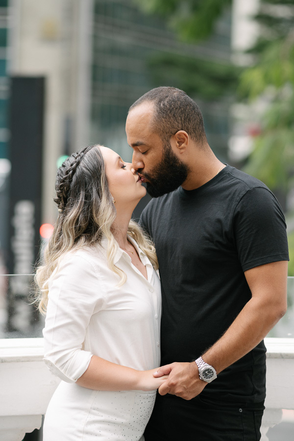 Claudia e Matheus se beijam carinhosamente em uma sacada na Avenida Paulista. Ela veste um conjunto branco e ele uma camiseta preta. Ao fundo, o cenário urbano de São Paulo com prédios modernos e árvores completa a atmosfera romântica do ensaio.