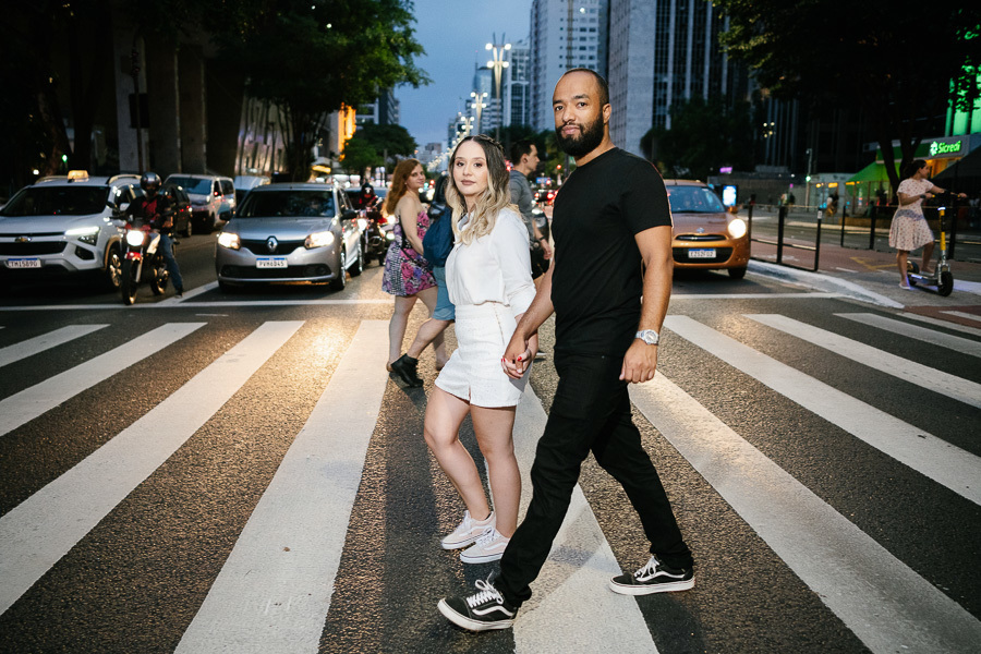 Claudia e Matheus atravessam de mãos dadas a faixa de pedestres da Avenida Paulista ao anoitecer. O casal veste branco e preto, destacando-se entre o movimento de carros e luzes da cidade. A cena captura a energia vibrante e urbana de São Paulo.