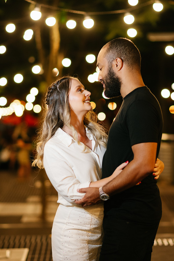 Claudia e Matheus trocam olhares apaixonados e sorrisos sob um varal de luzes decorativas ao ar livre à noite. Ela veste branco e ele preto. O fundo desfocado com as luzes amarelas circulares cria uma atmosfera mágica e acolhedora para o casal.