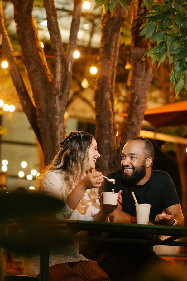 Claudia e Matheus sorriem e conversam enquanto tomam sorvete em uma mesa ao ar livre à noite. O ambiente é decorado com luzes amarelas suspensas entre as árvores, criando um clima alegre e romântico para encerrar o ensaio pré-wedding na Avenida Paulista.