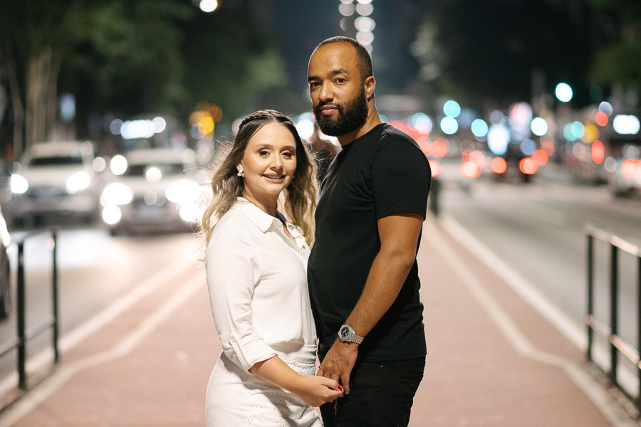 Claudia e Matheus posam de mãos dadas na ciclovia da Avenida Paulista à noite. Ela veste um conjunto branco e ele uma camiseta preta. Ao fundo, as luzes dos carros e da iluminação urbana criam um efeito bokeh, destacando o casal no centro da via.