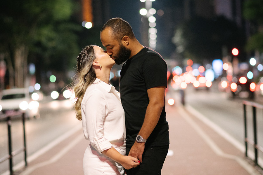 Claudia e Matheus se beijam apaixonados na ciclovia da Avenida Paulista durante a noite. Ela veste um conjunto branco e ele uma camiseta preta. Ao fundo, o brilho das luzes dos carros e da cidade cria um efeito bokeh romântico, iluminando o cenário urbano