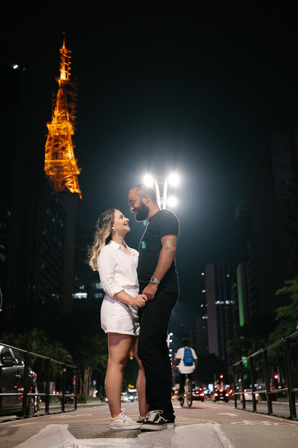 Claudia e Matheus trocam olhares apaixonados e sorrisos, de mãos dadas, na ciclovia da Avenida Paulista à noite. Ela veste um conjunto branco e ele uma camiseta preta. Ao fundo, a icônica antena iluminada de amarelo e as luzes dos prédios criam um cenário