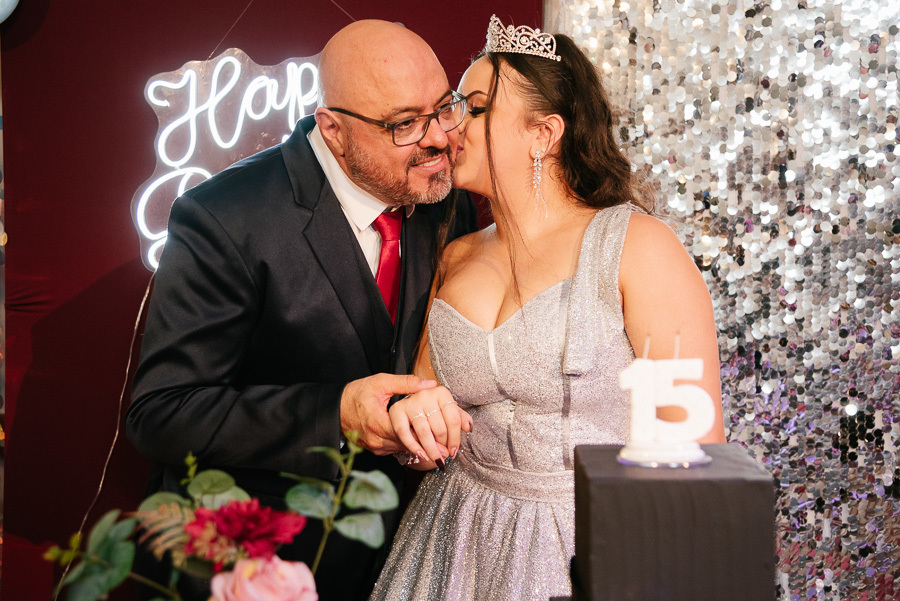 Amor em dobro: Debutante recebe um beijo carinhoso dos pais simultaneamente, foto clássica na mesa do bolo.