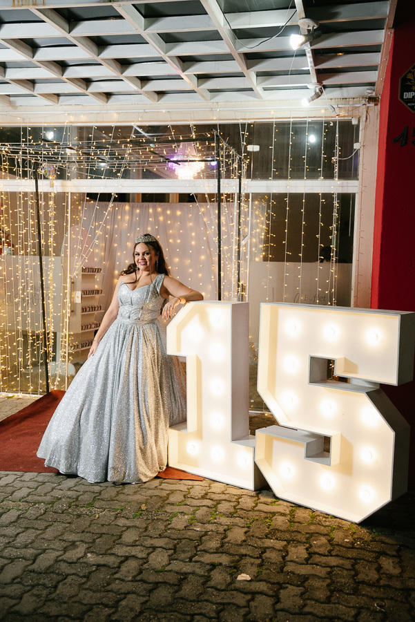 Poses descontraídas: Debutante sentada ao lado do letreiro 15 luminoso, aproveitando a decoração da festa.