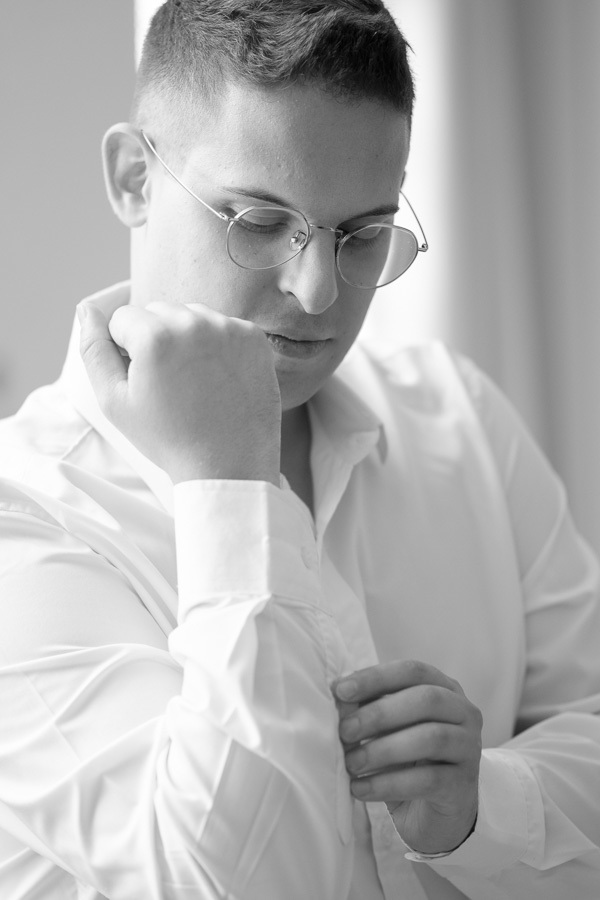 Making of do noivo Marcus em preto e branco. Detalhe artístico do noivo ajustando o punho da camisa na casa dos pais. Fotografia de casamento clássica e atemporal, capturando a concentração e a emoção antes da cerimônia.