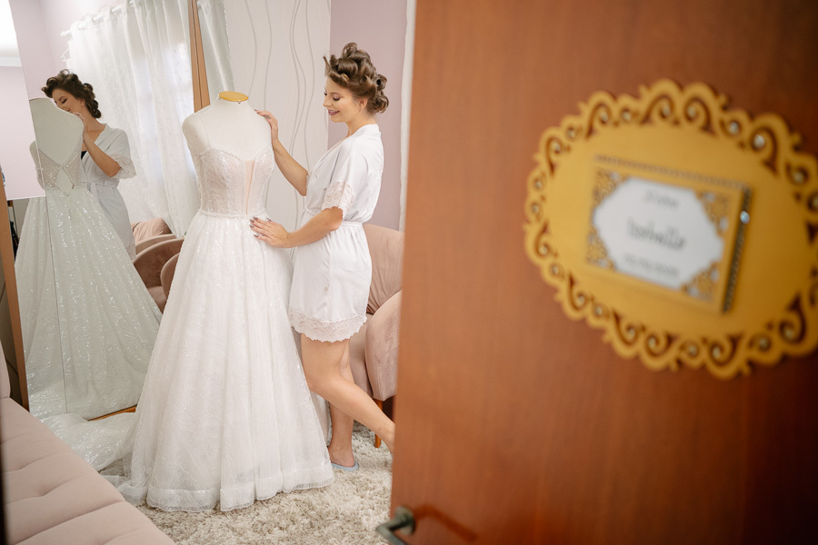 Noiva Isabelle admirando seu vestido de noiva momentos antes de se vestir. Composição criativa através da porta com a placa personalizada com seu nome. Making of emocionante registrado no C&D Hair e Makeup Studio.