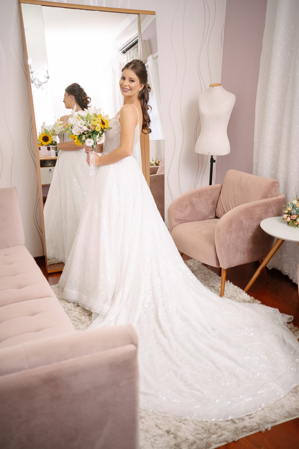 Noiva Isabelle pronta e deslumbrante em frente ao espelho. Foto de corpo inteiro mostrando a cauda do vestido de noiva estilo princesa e o buquê de girassóis. A elegância clássica antes de sair para a cerimônia.
