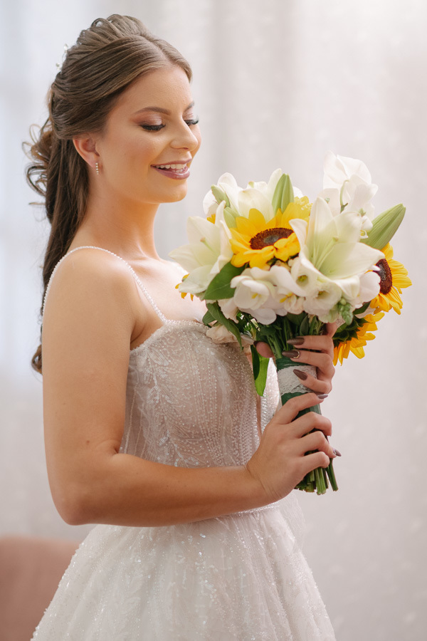 Close de perfil da noiva Isabelle sorrindo, segurando seu buquê de girassóis. Destaque para a maquiagem impecável, o penteado meio preso e os detalhes do corpete do vestido. Retrato romântico e iluminado.