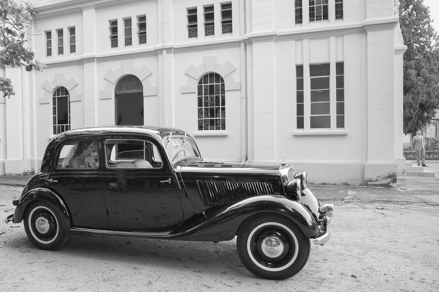 Chegada da noiva Isabelle em carro antigo clássico na Capela Sagrado Coração de Jesus, Parque Vicentina Aranha. Fotografia em preto e branco destacando a arquitetura histórica e a elegância do transporte vintage antes da cerimônia.
